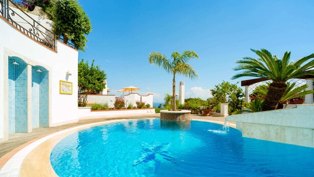 A swimming pool with a curved edge, surrounded by a white wall with blue-tiled windows, a palm tree, and a small fountain. - Villa Ambra by My Villa Collection (Casamicciola Terme)