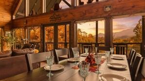 A dining area with a wooden table set for six, surrounded by chairs, and large windows offering a view of autumn foliage and mountains.