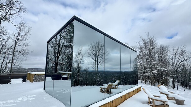 Een moderne glazen constructie met een houten terras en buiten zitplaatsen, omgeven door met sneeuw bedekte bomen en een bewolkte lucht.