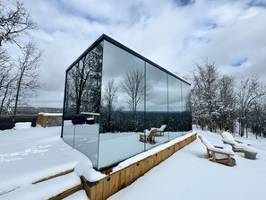 A modern glass structure with a wooden deck and outdoor seating, surrounded by snow-covered trees and a cloudy sky. - Romantic Couples Mirror Cabin | Hot Tub + Sauna (Faulkner County)