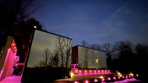 Een modern gebouw met een plat dak en grote ramen, omgeven door een houten terras met lichtslingers.