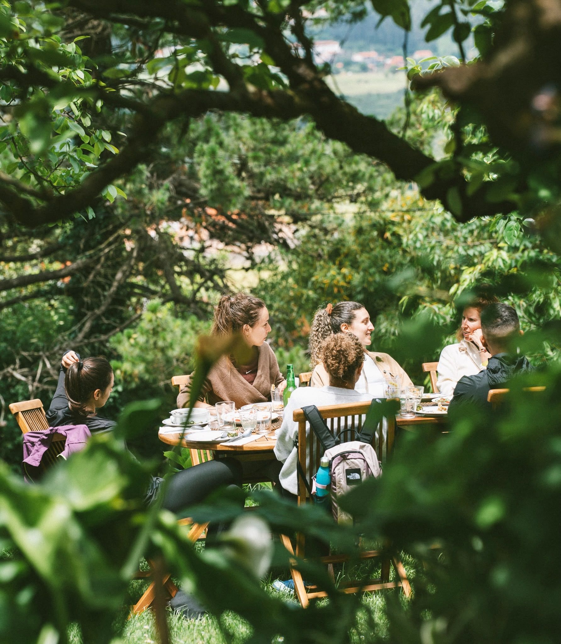 A group of people dining outdoors at a wooden table surrounded by greenery.
