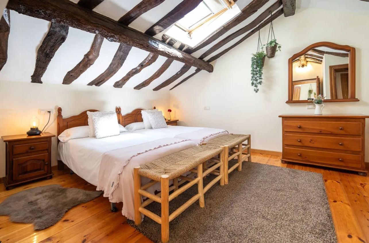 Une chambre avec un plafond en bois, un lit avec une literie blanche, un banc, une commode et un miroir.