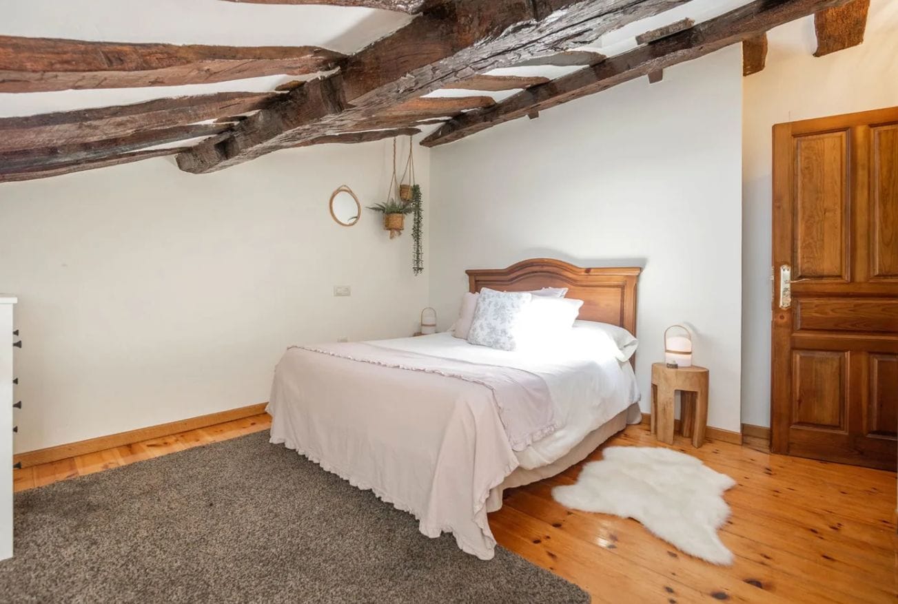 A bedroom with a wooden ceiling, a bed with white bedding, a small bedside table, and a door.