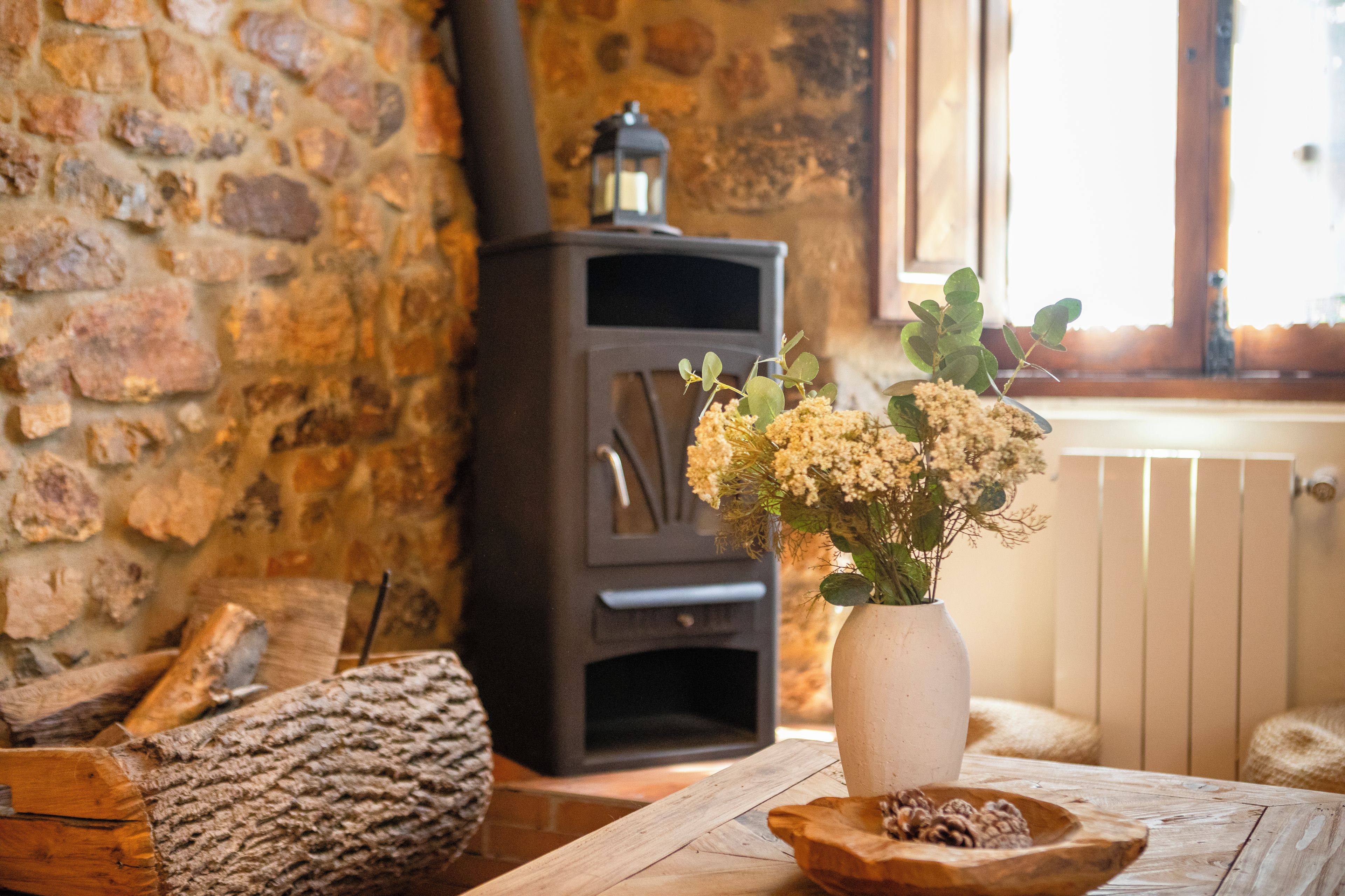 A rustic interior with a stone wall, a wood-burning stove, a vase with flowers, and a wooden table.