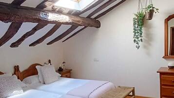 A bedroom with a sloped ceiling, wooden beams, a bed with white linens, a bench, and a hanging plant.