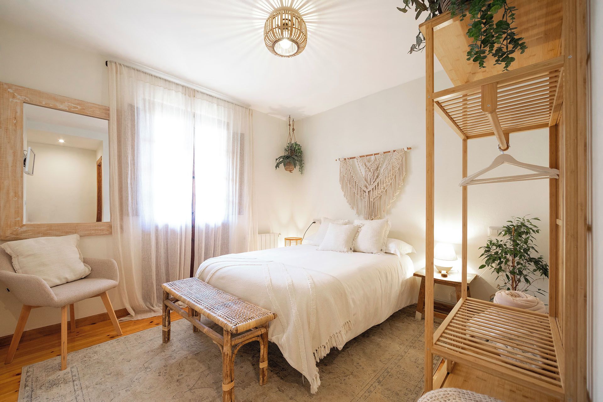 Une chambre à coucher comprenant un lit, une chaise, un banc, une décoration murale en macramé suspendue et une étagère en bois avec des plantes.