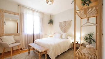 A bedroom with a bed, a chair, a bench, a hanging macrame wall hanging, and a wooden shelf with plants.