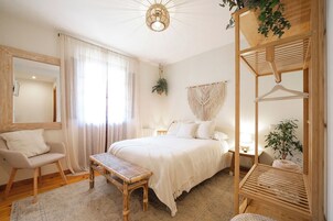 A bedroom with a bed, a chair, a bench, a hanging macrame wall hanging, and a wooden shelf with plants.
