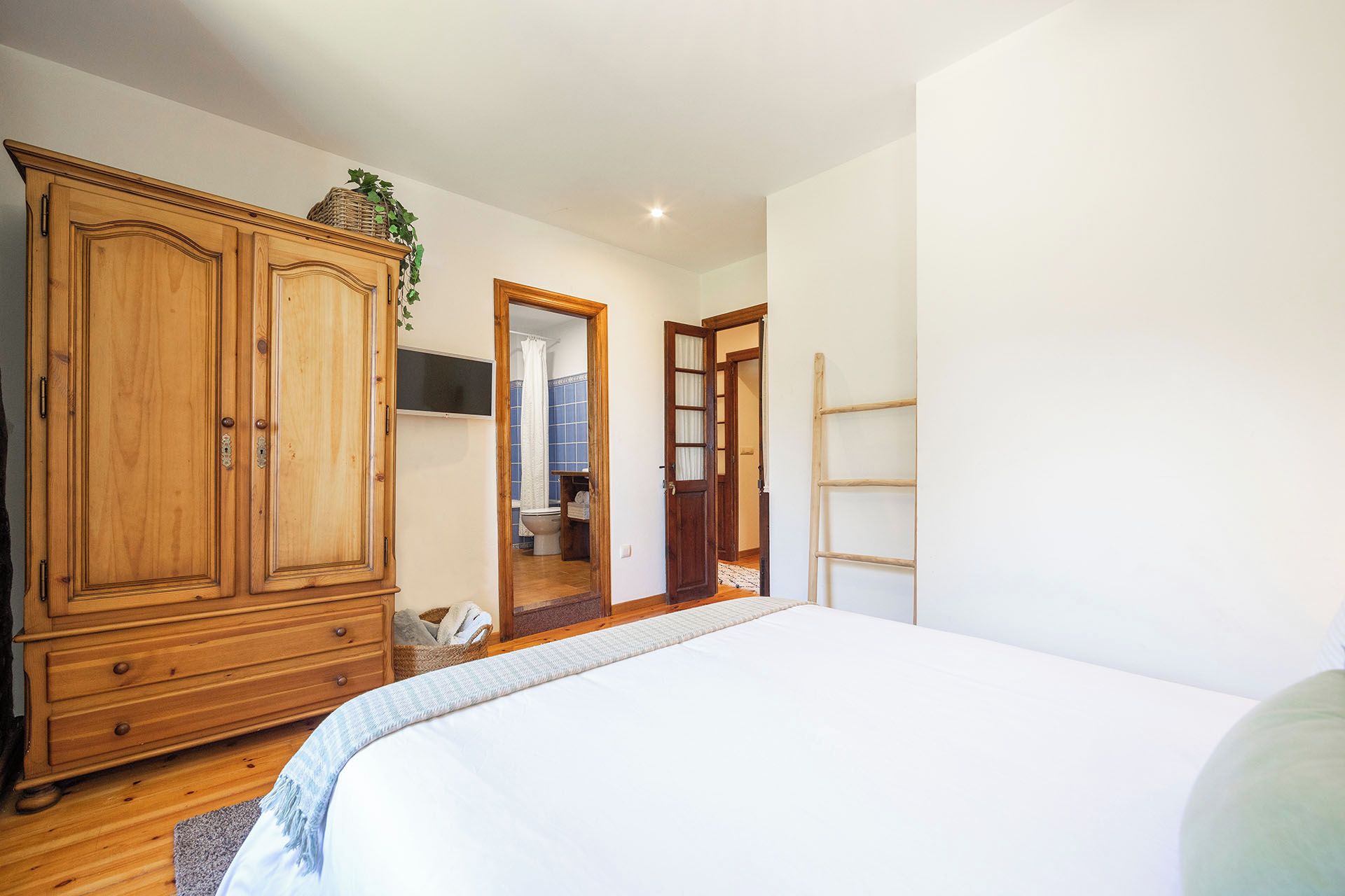 Un dormitorio con un armario grande de madera, una cama con edredón blanco y un baño visible a través de una puerta abierta.