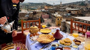 Una mesa servida con platos tradicionales marroquíes, un juego de té y vasos, con vistas a un paisaje urbano con edificios históricos.