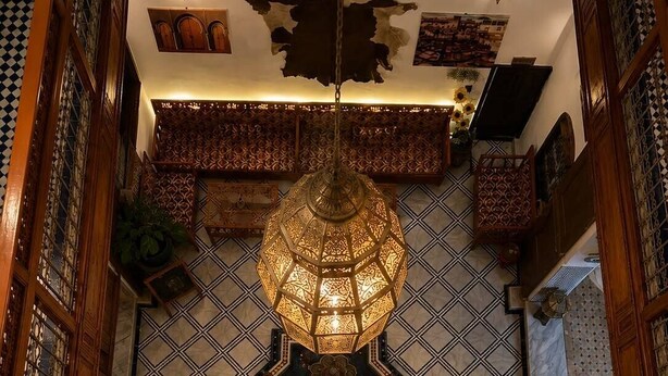 A room with a patterned floor, a hanging lantern, and a wall-mounted tapestry. - Riad hamad (Fes)