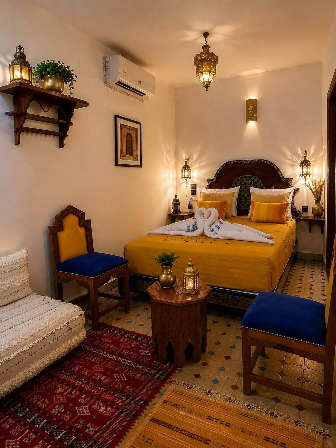 A hotel room with a bed, two chairs, a small table, a decorative lamp, and a wall-mounted air conditioner.