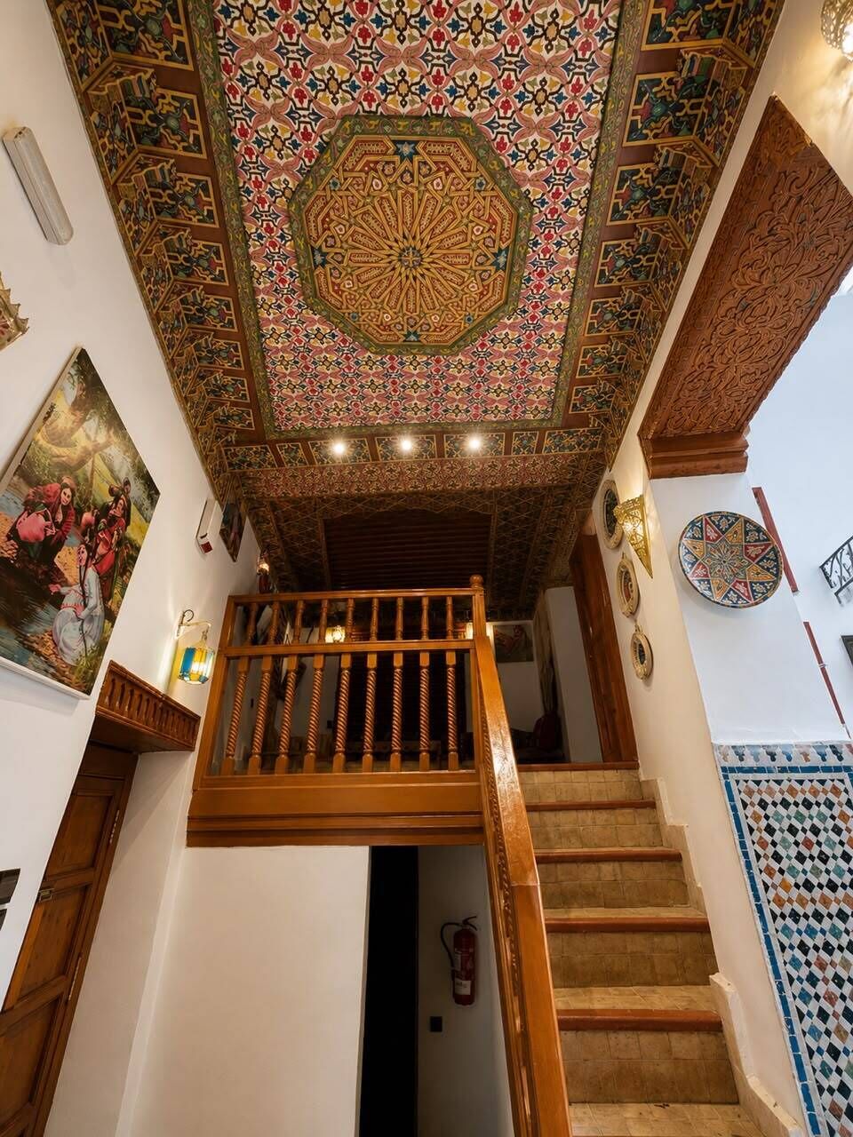 Escadaria com corrimão de madeira que leva a um patamar, cercada por paredes com desenhos elaborados em azulejos e uma grande moldura decorativa no teto.