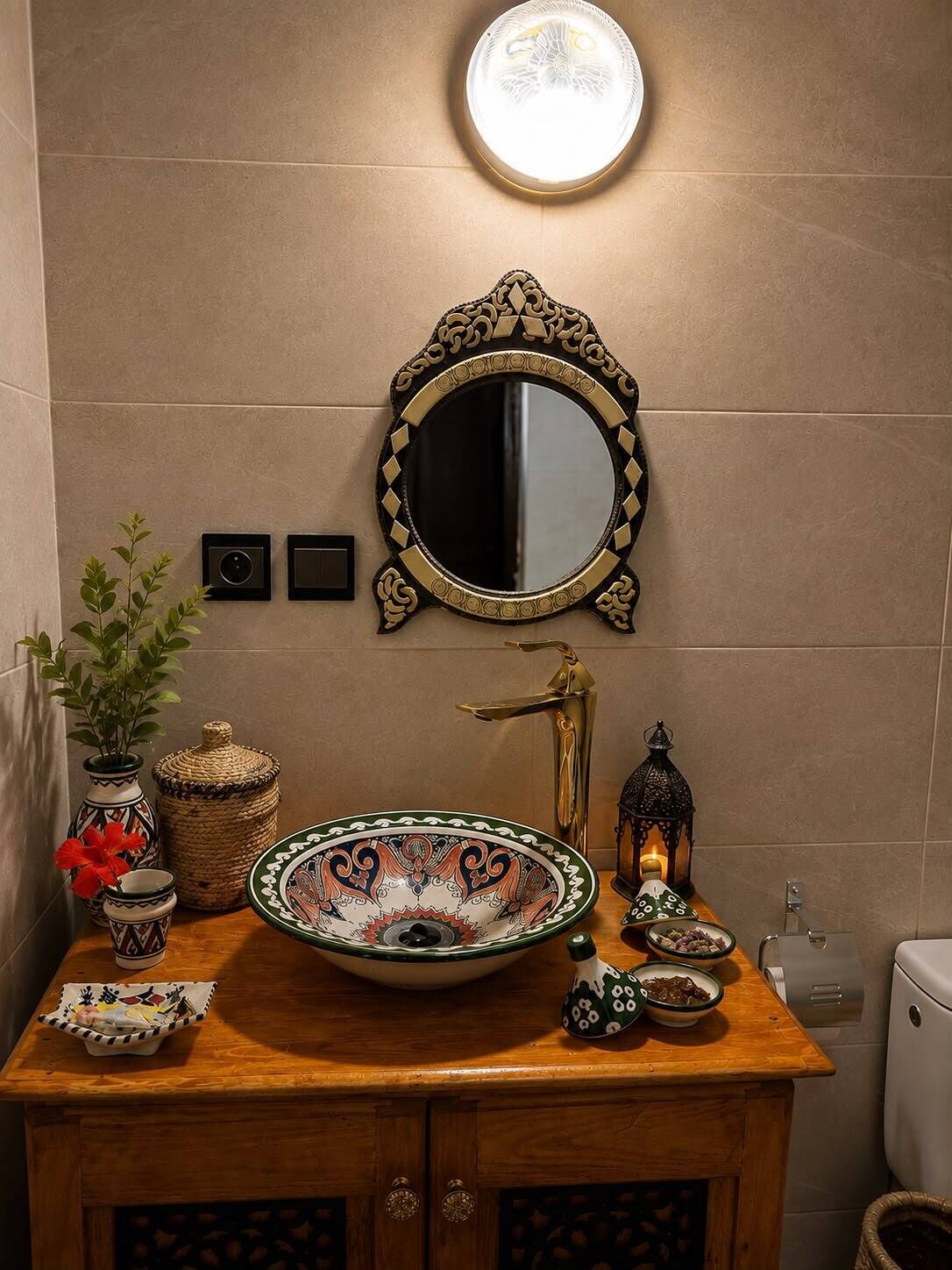 Een badkamer met een ronde spiegel, een wastafel op voet, een decoratieve vaas, een klein plantje, een kaars en een lantaarn.