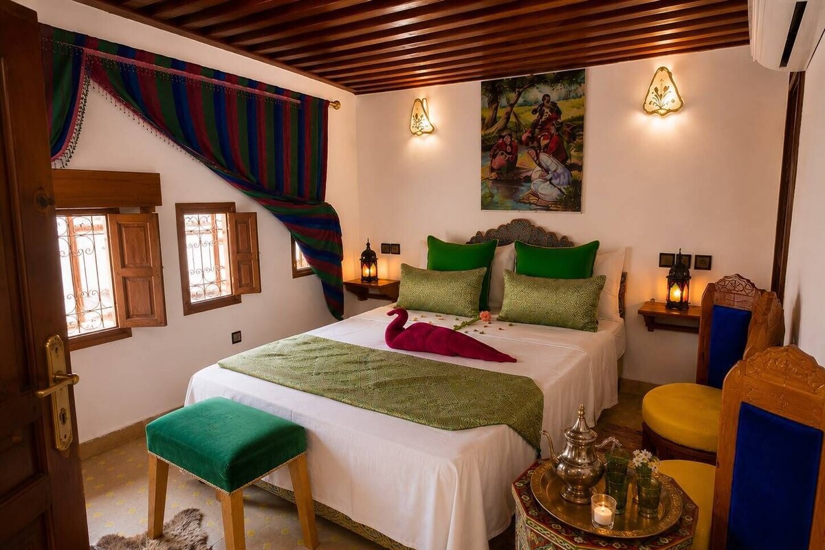 A bedroom with a bed, a bench, a table with a lamp, and a painting on the wall.