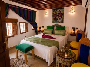 A bedroom with a bed, a bench, a table with a lamp, and a painting on the wall. - Riad hamad (Fes)