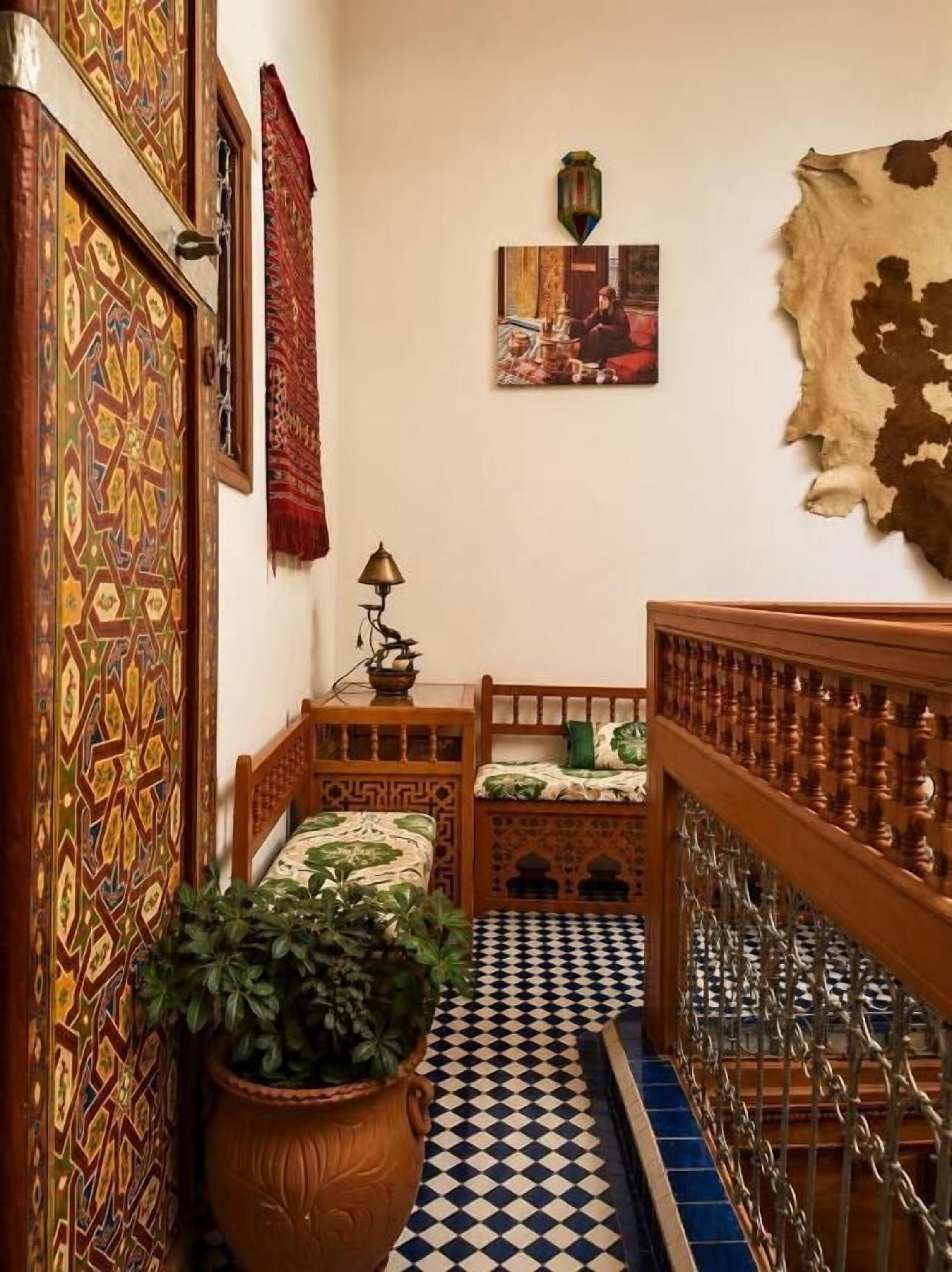 Un pasillo estrecho con paredes estampadas, piso a cuadros, cama con cubrecama verde, planta en maceta y un tapiz colgado.
