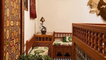 Un pasillo estrecho con paredes estampadas, piso a cuadros, cama con cubrecama verde, planta en maceta y un tapiz colgado.