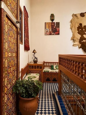 A narrow hallway with patterned walls, a checkered floor, a bed with a green bedspread, a potted plant, and a hanging tapestry. - Riad hamad (Fes)