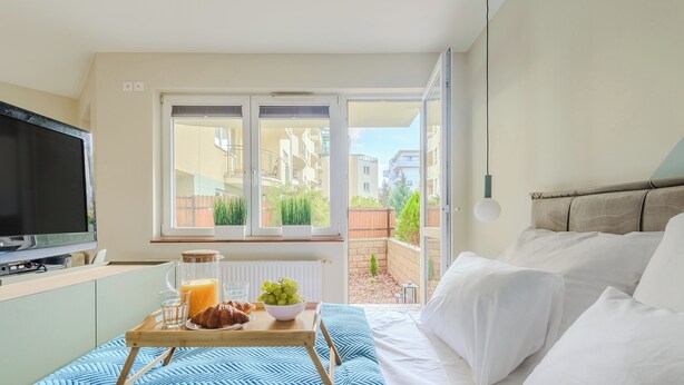 Quarto com cama, bandeja de café da manhã com croissants e frutas, televisão e janela com vista para um jardim e edifícios.
