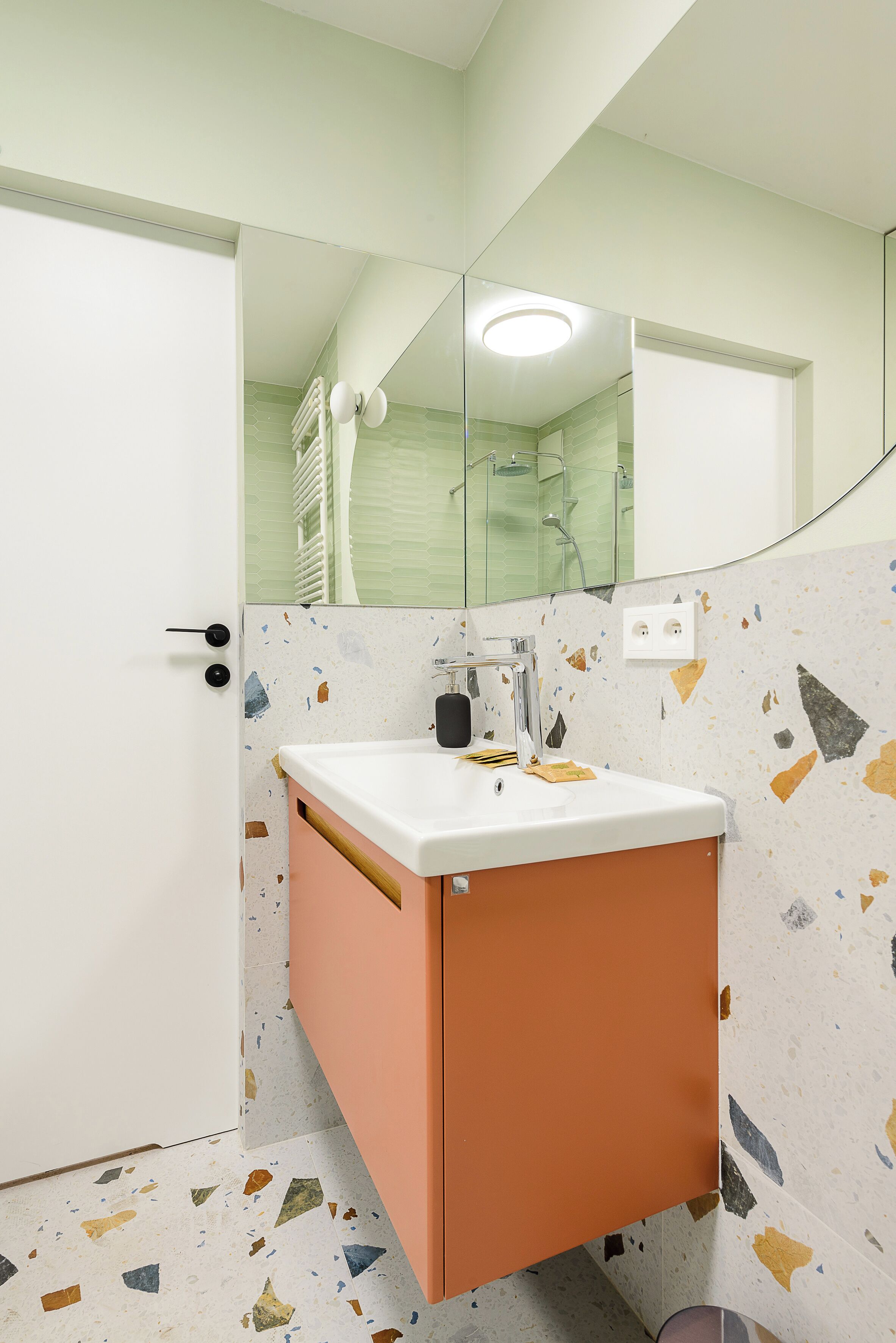 Een badkamer met een witte deur, een wastafel met een spiegel erboven en een zichtbare douchecel in de weerspiegeling.