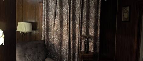 A living room with a dark wood-paneled wall, a patterned curtain, a lamp, a chair, and a small table with a vase.