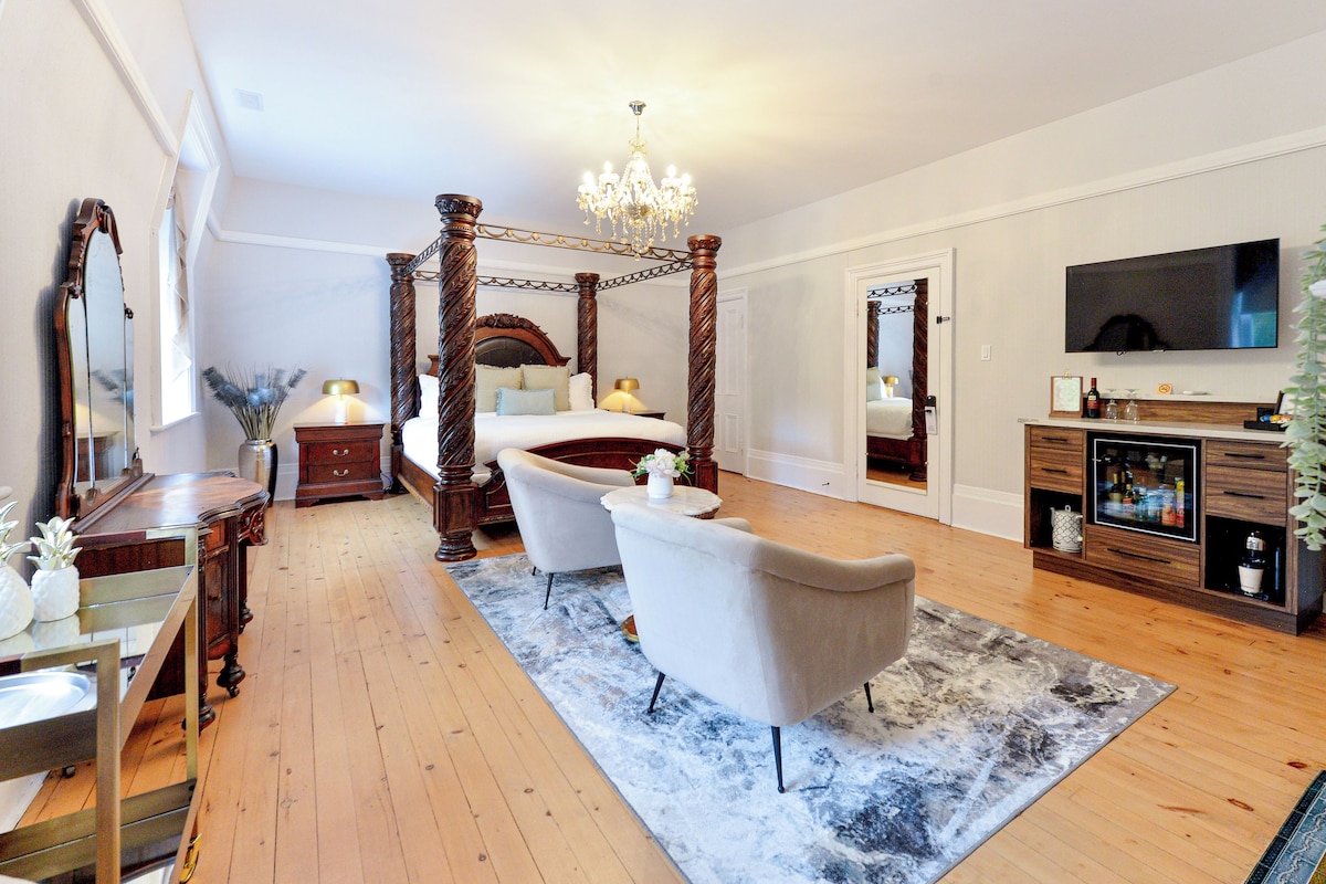 A spacious bedroom with a four-poster bed, a sitting area with a chair, a vanity with a mirror, and a television on the wall.