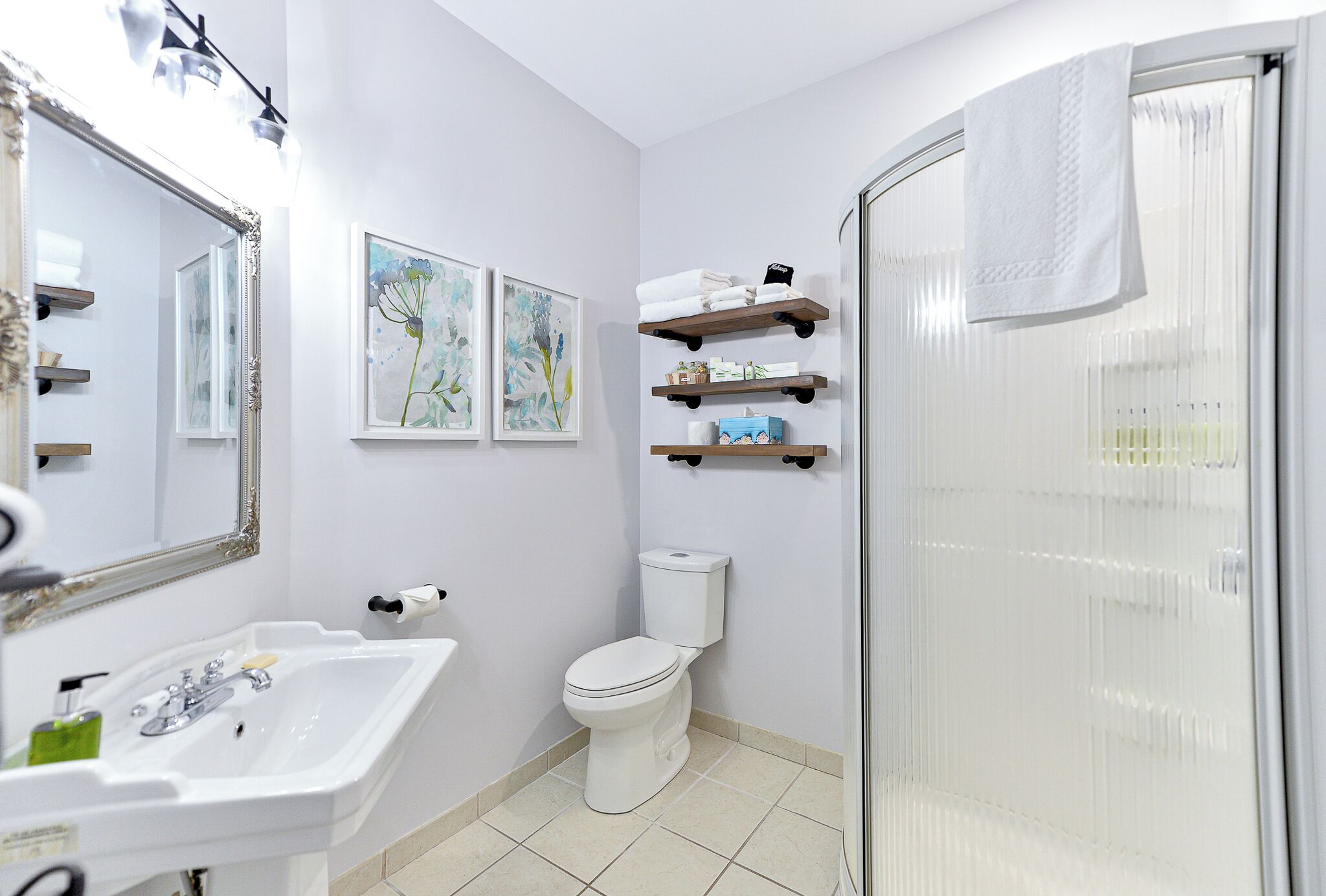 A bathroom with a white sink, toilet, and shower. There are three shelves above the toilet with toiletries.