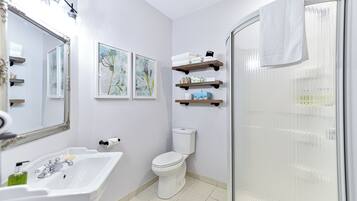 A bathroom with a white sink, toilet, and shower. There are three shelves above the toilet with toiletries.