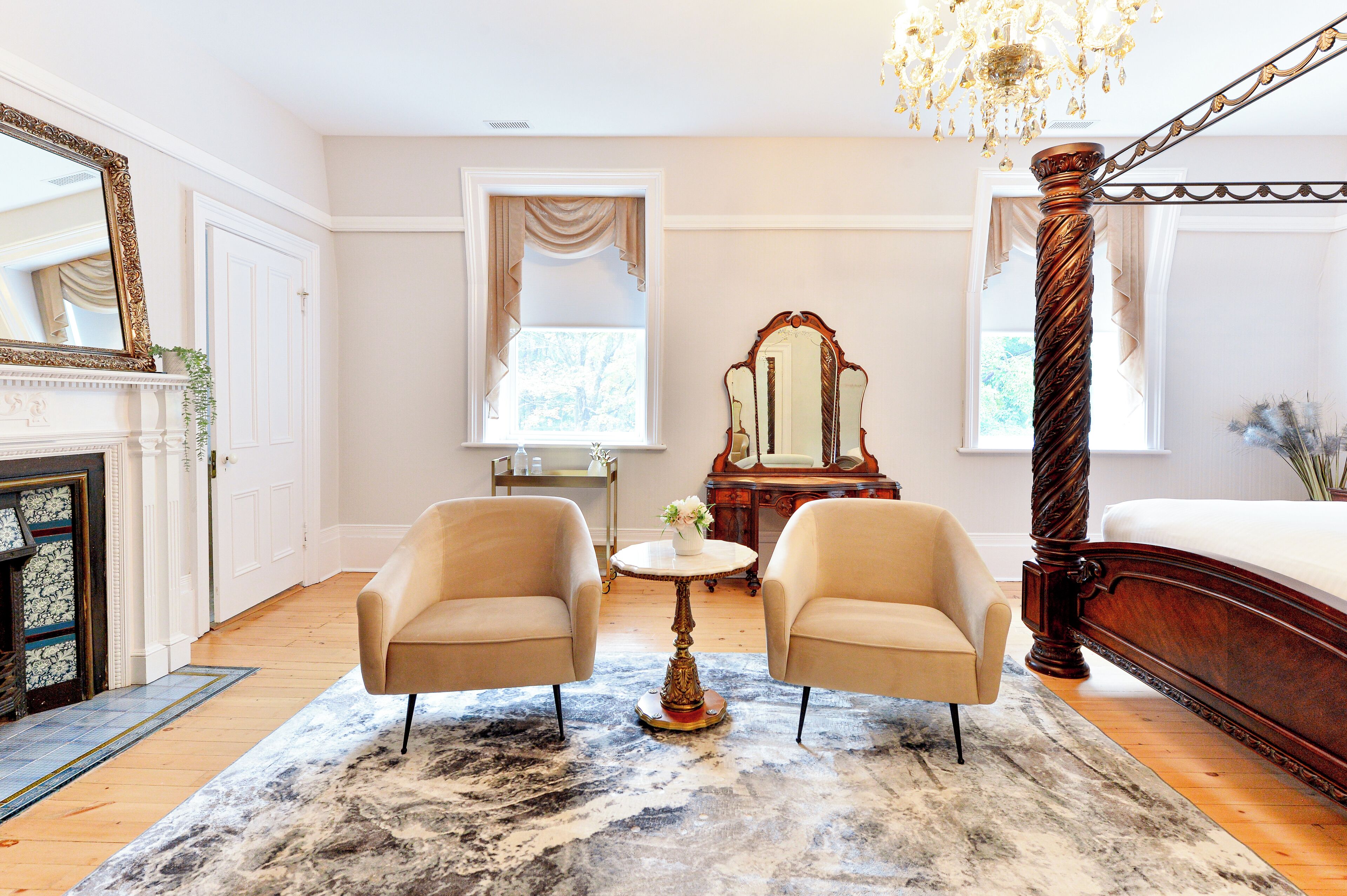 A bedroom with a large bed, two armchairs, a round table, a mirror, and a chandelier.