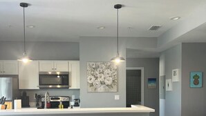 Una cucina moderna con mobili bianchi, un'isola centrale con sgabelli da bar, illuminazione a sospensione e un grande quadro floreale appeso al muro.