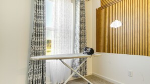 Une pièce d’angle avec un mur lambrissé en bois, une fenêtre ornée de rideaux légers et un tableau à repasser pliable.