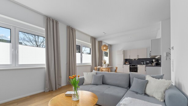 A modern living room with a grey sofa, wooden coffee table, and a vase of flowers. A kitchen is visible in the background with white cabinets and a black countertop. - Central | Modern | Old Town | Parking lot | 70 m² (Bad Kreuznach)