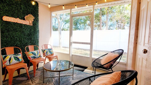 Una habitación con una mesa de vidrio, dos sillas naranjas con cojines de colores, una silla de mimbre negra y una pared con un tapiz verde y un cartel.