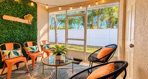 Un patio interior con mesa de vidrio, sillas de mimbre naranja y negro, y una pared verde con decoración de palmera.