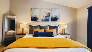 Habitación de hotel con una cama grande, dos mesitas de noche con lámparas, un espejo y tres cuadros abstractos en la pared.