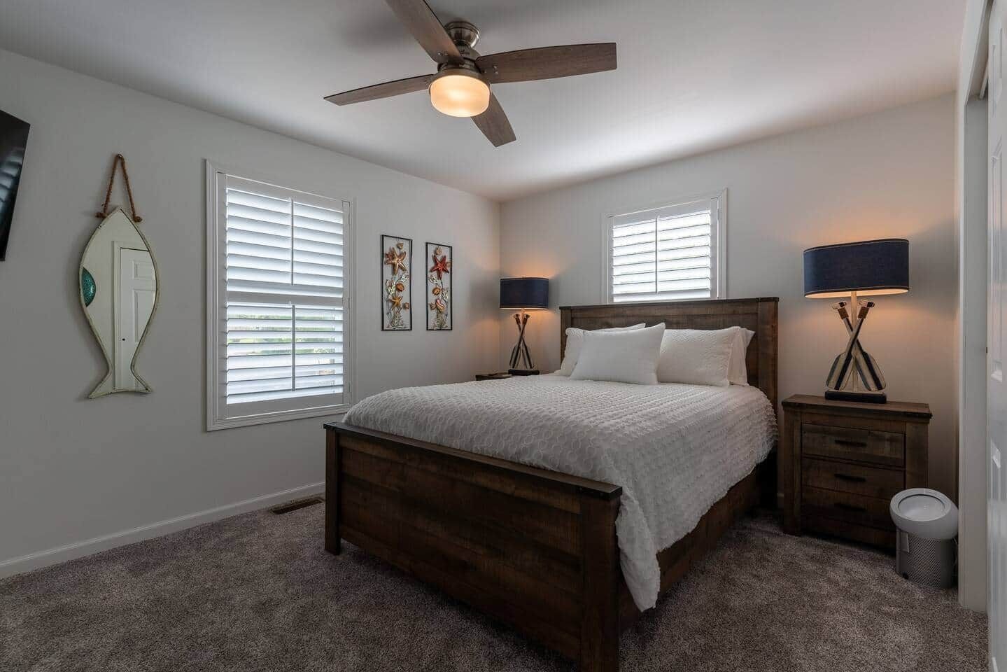 A bedroom with a wooden bed, two bedside tables with lamps, a ceiling fan, and two windows with blinds.