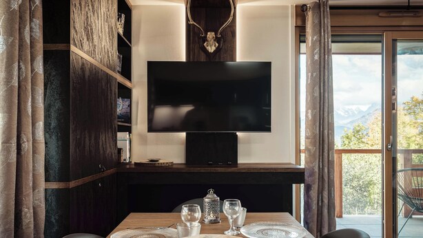 A dining area with a wooden table, chairs, and a television mounted on a dark wall.