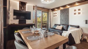 A modern living room with a wooden dining table set for four, a black leather sofa, and a flat-screen TV mounted on the wall.