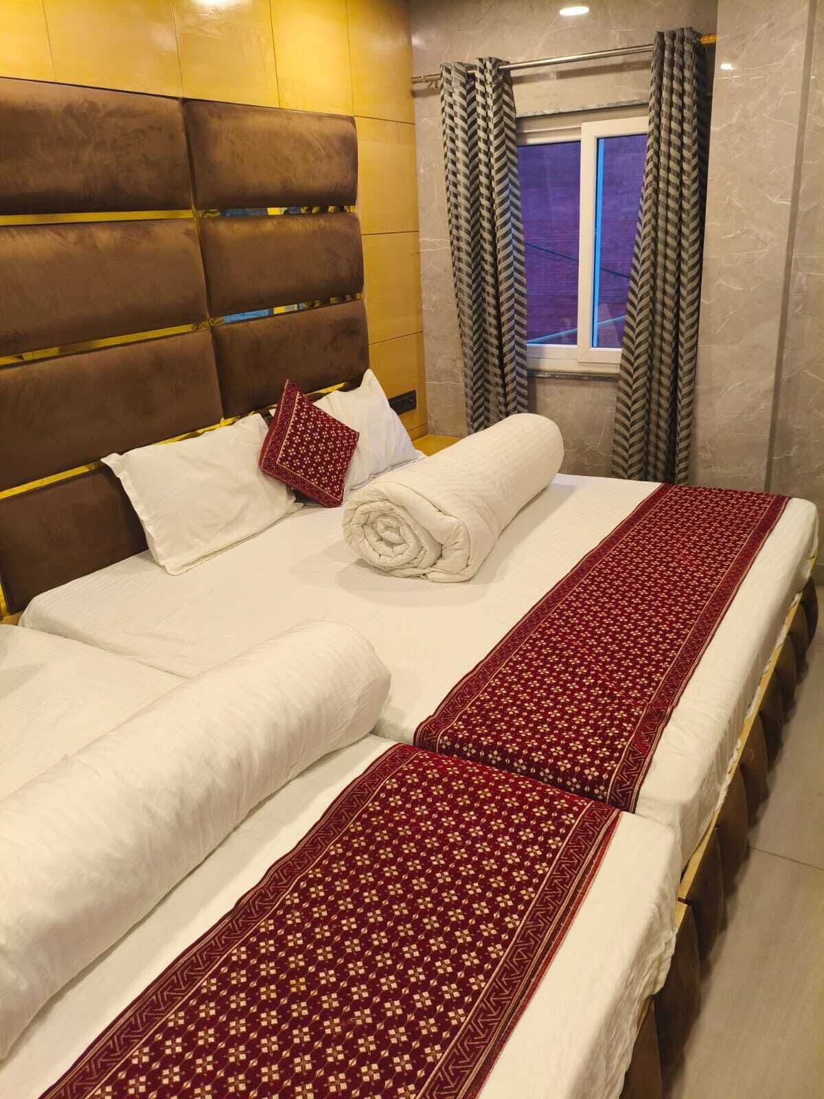 A neatly made bed with white linens and a red patterned bedspread, a brown headboard, and a window with curtains.