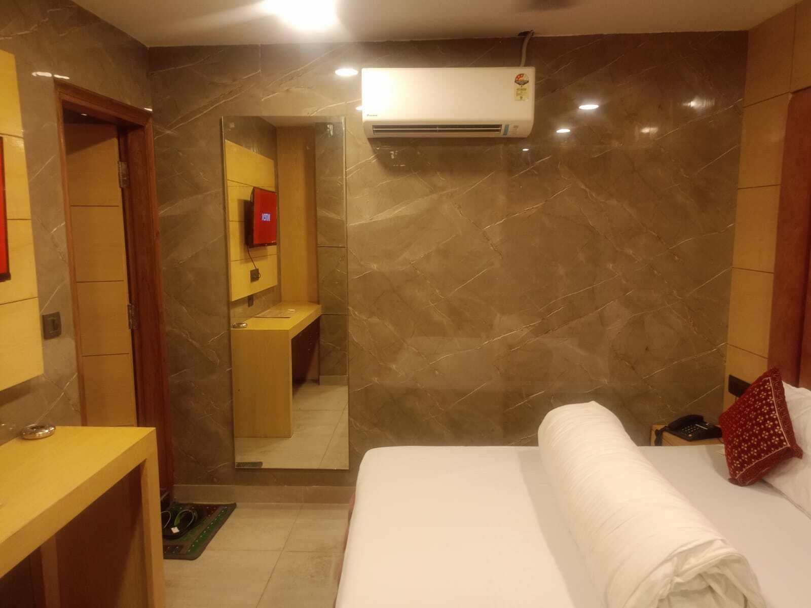 A hotel room with a bed, a desk, a mirror, and an air conditioning unit.