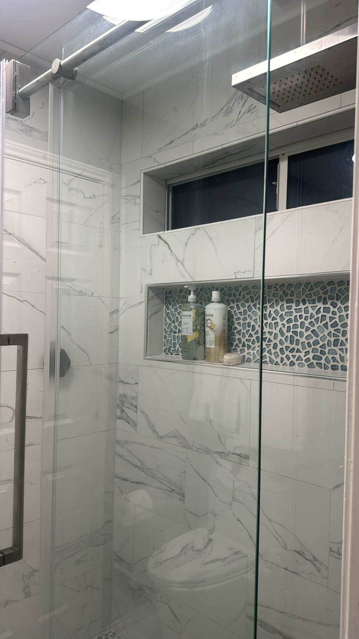 A bathroom with a glass shower enclosure, marble tiles, and a shelf with toiletries.