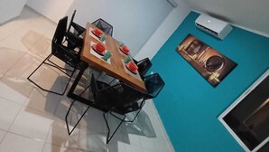 A dining area with a table set for four, surrounded by black chairs. There is a ceiling light, a wall-mounted air conditioner, and a framed picture on the wall.