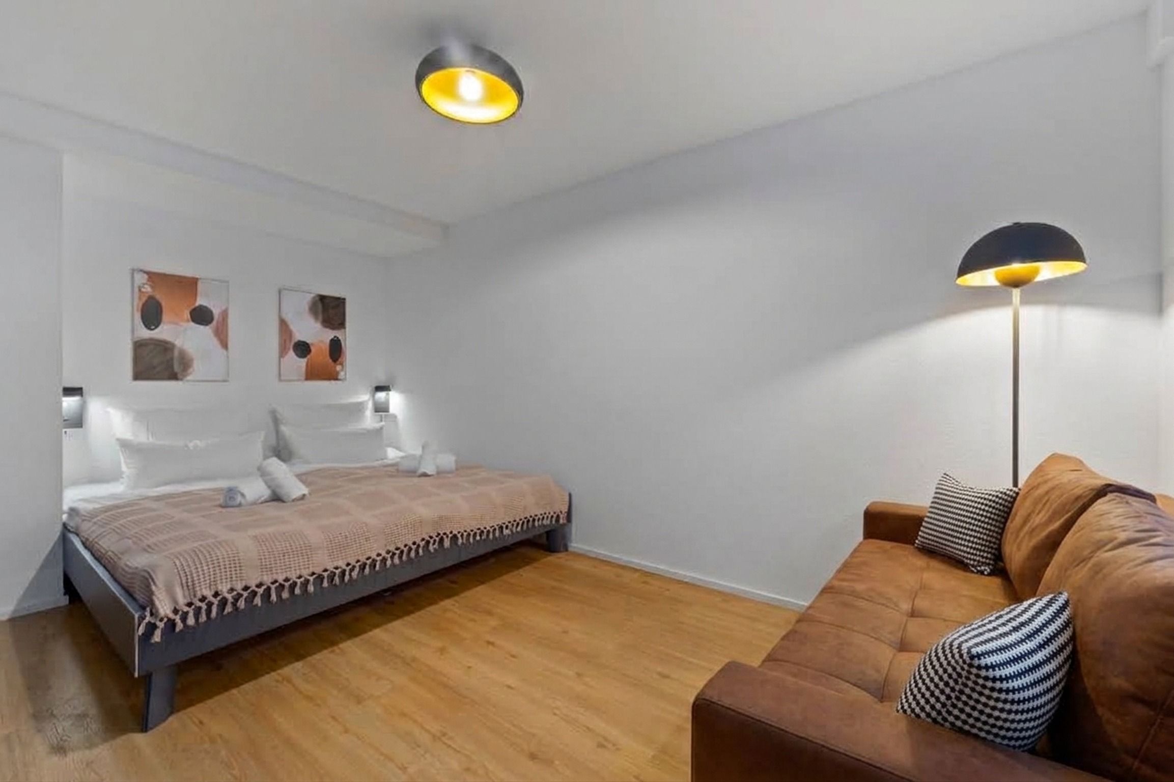 Une chambre à coucher avec un lit, un canapé marron, une lampe sur pied et des œuvres d’art abstraites accrochées au mur.