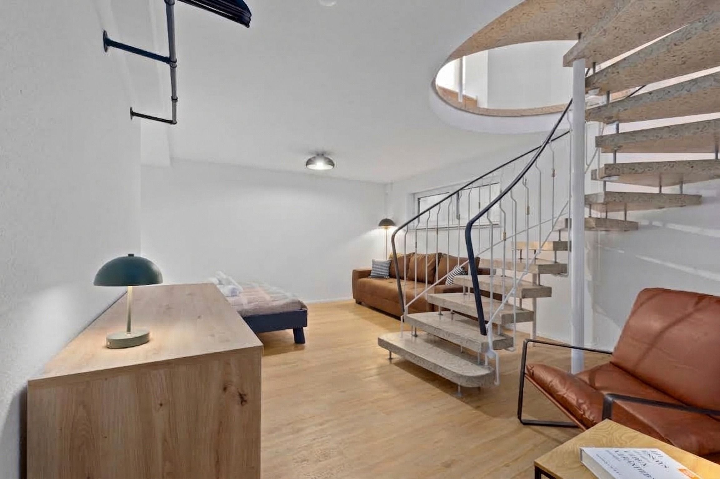 A modern interior with a wooden desk, a sofa, a brown leather chair, and a spiral staircase.