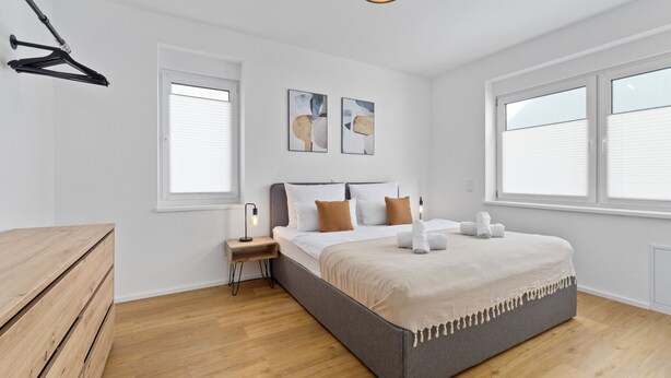 A bedroom with a bed, bedside tables, a hanging light fixture, and two windows. - Maisonette | Old Town | Parking lot | 96 m² | Modern (Bad Kreuznach)