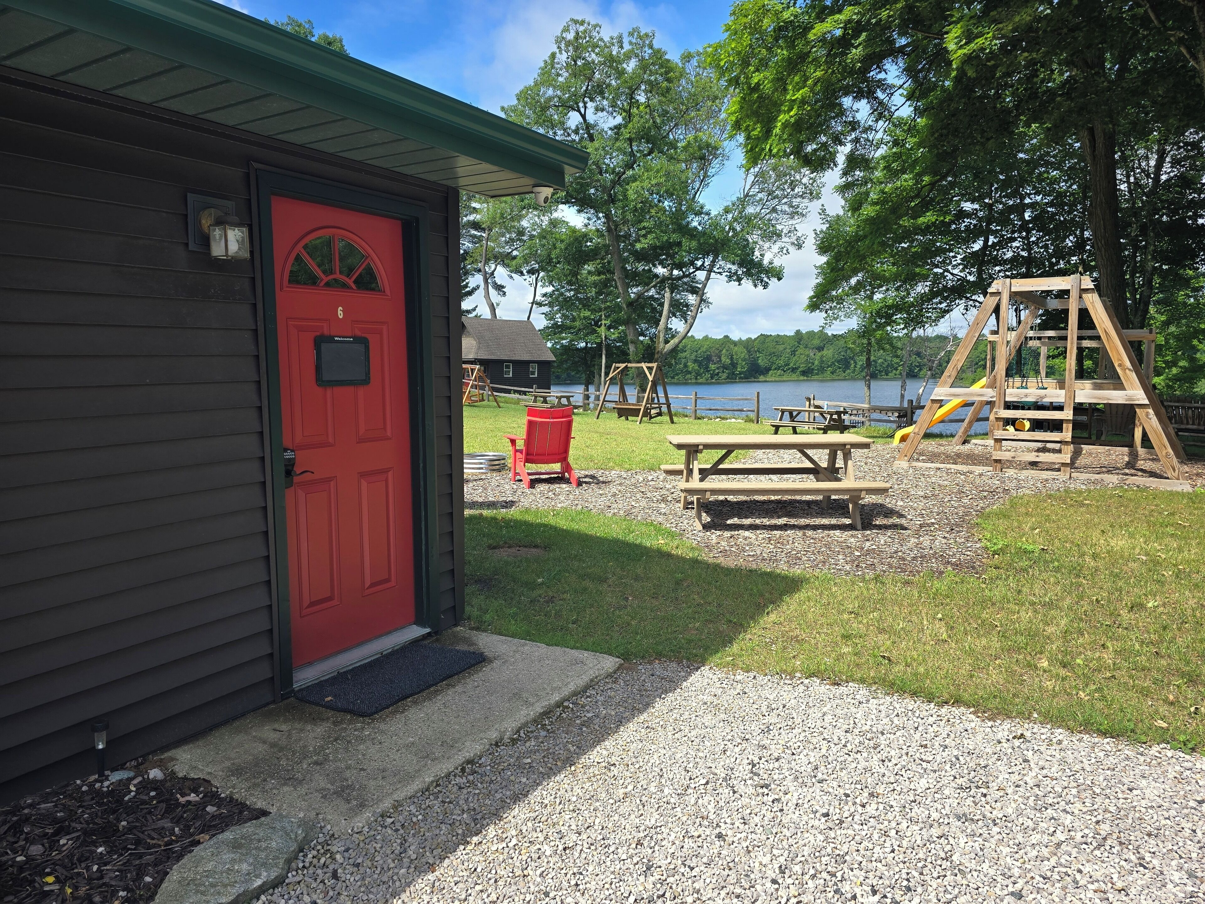 A cabin with a red door, a picnic table, and a swing set in a park with a lake in the background.