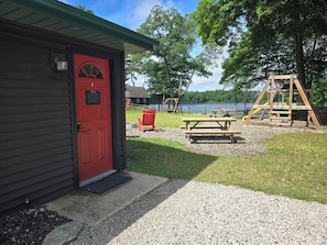 A cabin with a red door, a picnic table, and a swing set in a park with a lake in the background. - Sleeping Bear Resort (Lake Ann)