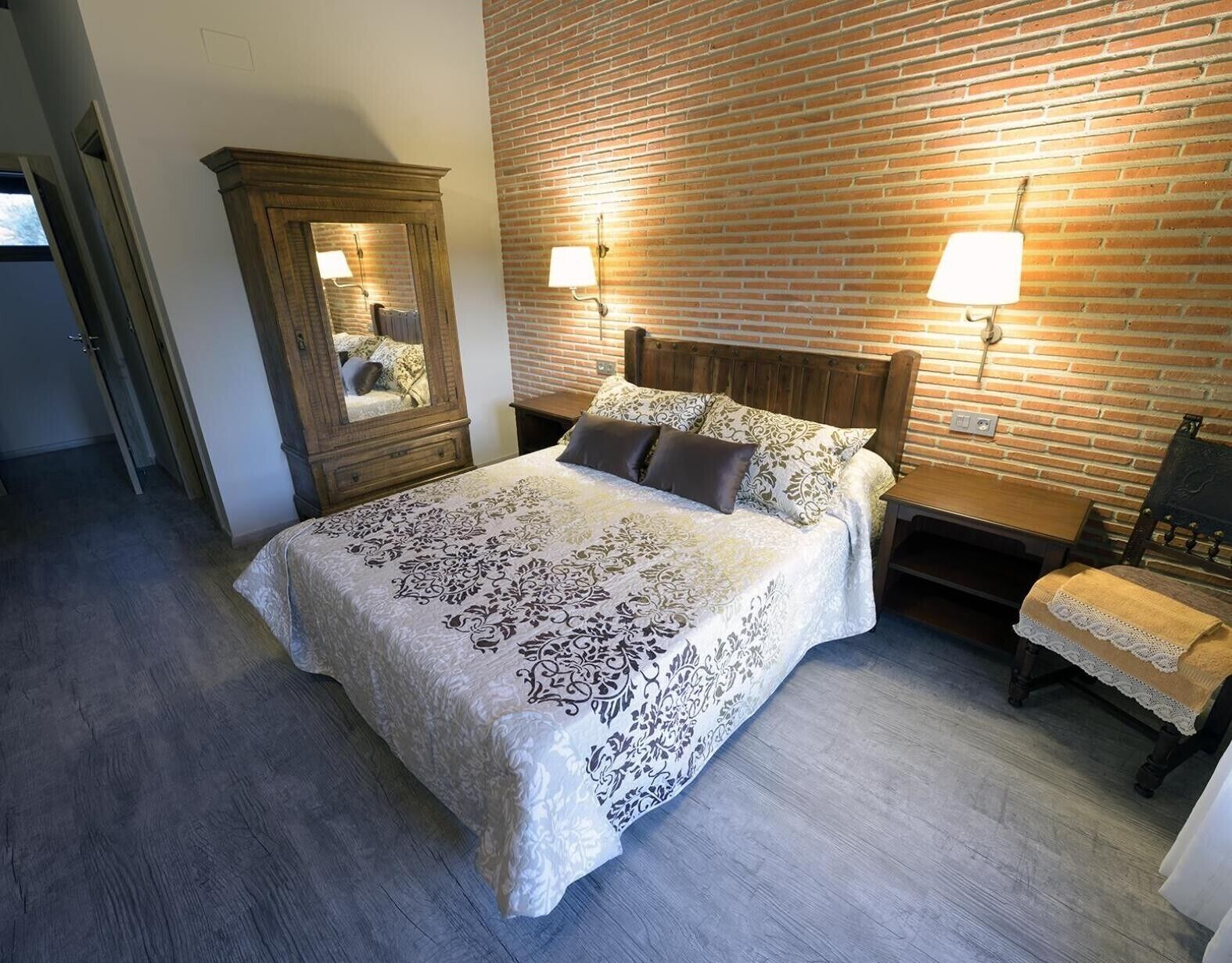 A bedroom with a large bed, a wooden wardrobe, a nightstand, a chair, and a brick wall.